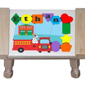 Puede incluir: Un taburete de madera con una tapa de rompecabezas colorida que presenta el nombre "Ethan", un camión de bomberos, una estación de bomberos y varias formas geométricas. El taburete tiene patas torneadas.