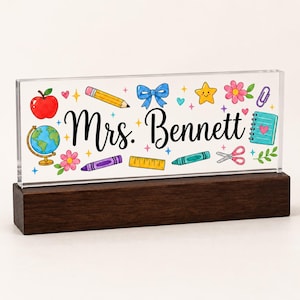 May include: A clear acrylic desk sign with a dark wood base featuring the text Mrs. Bennett in black script. The design includes colorful illustrations of a red apple, a globe, a yellow pencil, a blue bow, a star, flowers, a ruler, and scissors.