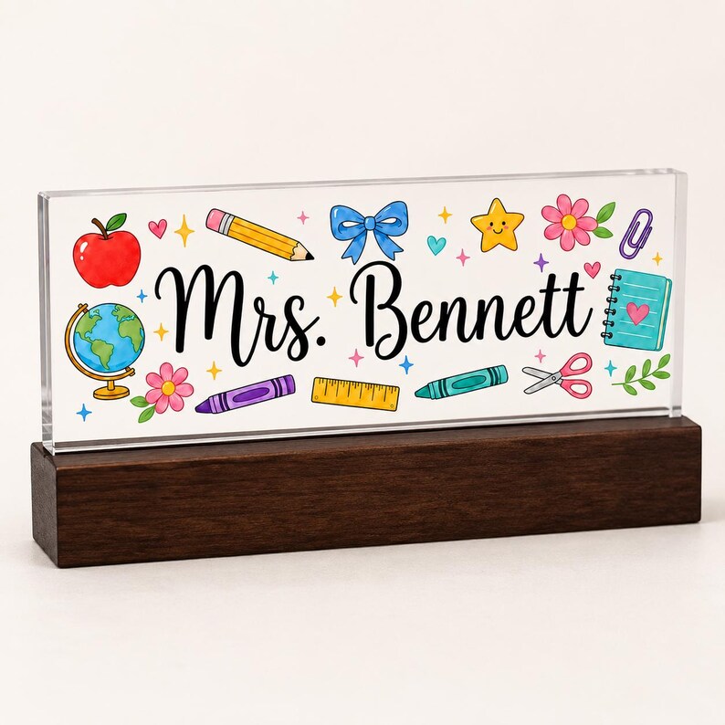May include: A clear acrylic desk sign with a dark wood base featuring the text Mrs. Bennett in black script. The design includes colorful illustrations of a red apple, a globe, a yellow pencil, a blue bow, a star, flowers, a ruler, and scissors.