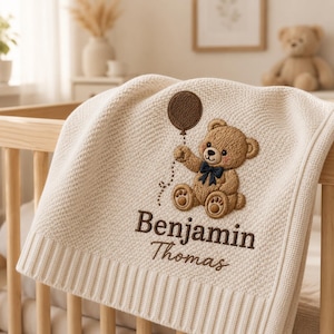 May include: A cream-colored knit baby blanket featuring an embroidered brown teddy bear holding a dark brown balloon. The bear wears a navy blue bow tie. Below the bear, the name Benjamin Thomas is embroidered in dark brown, serif lettering.