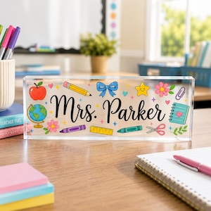 May include: A rectangular clear acrylic desk sign featuring the black script text Mrs. Parker. The design includes colorful illustrations of a red apple, a globe, a yellow pencil, a blue bow, a star, flowers, a ruler, scissors, and a notebook.