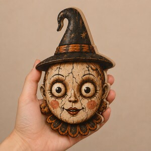May include: A flat, layered wooden cutout shaped like a doll head wearing a pointed witch hat. The face has a cracked texture, large round eyes, rosy cheeks, and a ruffled collar. The color palette includes black, tan, and orange accents.