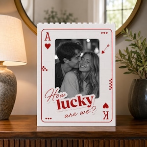 May include: A white rectangular photo frame constructed from interlocking building blocks, designed to resemble a playing card. It features red heart symbols, the letter A, and the text How lucky are we? in red, framing a black and white photograph.
