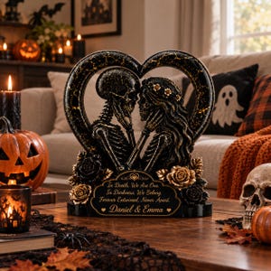 May include: A black and gold heart-shaped sculpture featuring two skeletons facing each other, adorned with roses. The base displays the text: In Death, We Are One. In Darkness, We Belong. Forever Entwined, Never Apart. Daniel & Emma.
