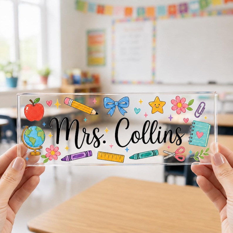 May include: A clear rectangular desk sign featuring the black script text Mrs. Collins. The design includes colorful illustrations of a red apple, a globe, a yellow pencil, a blue bow, a yellow star, a pink flower, a paperclip, a notebook, a ruler, a purple crayon, a green crayon, and pink scissors.