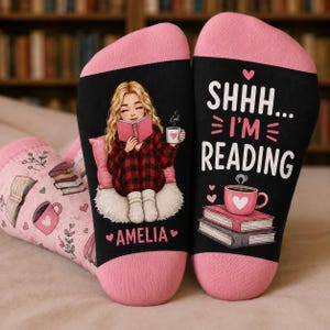 May include: A pair of black and pink socks. One sock features a blonde character reading a book, wearing a red and black plaid shirt, with the name AMELIA printed below. The other sock displays the text SHHH... I'M READING above a stack of books and a mug.