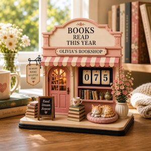 May include: A miniature bookshop display featuring a pink and cream facade with the text BOOKS READ THIS YEAR and OLIVIA'S BOOKSHOP. It includes a hanging sign reading SO MANY BOOKS SO LITTLE TIME, a chalkboard sign reading Read Dream Repeat, a flip counter showing 075, stacks of books, a sleeping orange tabby cat, and a potted plant.