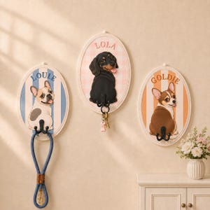 Op de afbeelding: Drie ovale wandhaken met illustraties van honden. De linkerhaak toont een Franse Bulldog met het label LOULLE op een blauw gestreepte achtergrond. De middelste haak toont een teckel met het label LOLA op een roze ruitjesachtergrond. De rechterhaak toont een Chihuahua met het label GOLDIE op een oranje gestreepte achtergrond.