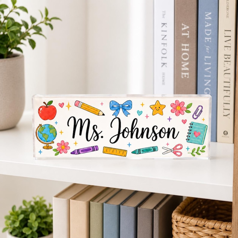 May include: A rectangular clear acrylic desk sign featuring the black text Ms. Johnson in the center, surrounded by colorful illustrations of a red apple, a globe, pencils, a blue bow, a yellow star, flowers, a ruler, and school supplies.