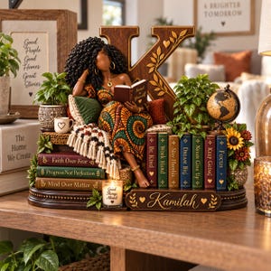 Puede incluir: Una figura decorativa que muestra a una persona con cabello oscuro rizado leyendo un libro titulado Invest in Yourself, sentada contra una letra K grande. La base incluye libros con frases motivadoras, un globo terráqueo y el nombre Kamilah.