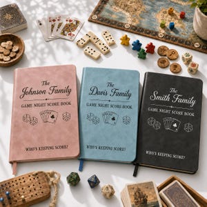 Puede incluir: Tres libros de puntuación rectangulares en rosa, azul y negro, cada uno con el texto 'Game Night Score Book', nombres de familia personalizados, dados e ilustraciones de cartas. Las portadas incluyen la frase 'Who's Keeping Score?' y un marcapáginas.