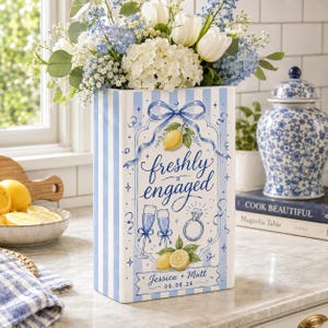May include: A rectangular ceramic vase with light blue and white vertical stripes. The front features a blue illustration of a bow, lemons, two champagne flutes, and a diamond ring with the text "freshly engaged" and "Jessica & Matt 06.08.26".