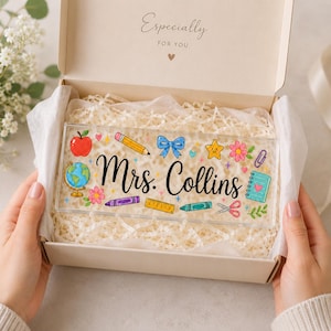 May include: A rectangular clear acrylic desk sign featuring the black script text Mrs. Collins, surrounded by colorful illustrations of a globe, apple, pencil, blue bow, star, flower, ruler, crayon, scissors, and a notebook inside a gift box.