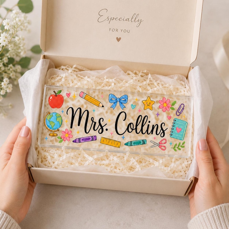 May include: A rectangular clear acrylic desk sign featuring the black script text Mrs. Collins, surrounded by colorful illustrations of a globe, apple, pencil, blue bow, star, flower, ruler, crayon, scissors, and a notebook inside a gift box.