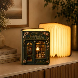 Op de afbeelding: Een donkergroen boekenhoek-diorama met de tekst Eliana's Book Nook, voorzien van een miniatuur winkelpui met een boogdeur, een verlicht raam en bloemaccenten. Daarnaast staat een boekvormige lamp die een warm schijnsel verspreidt.