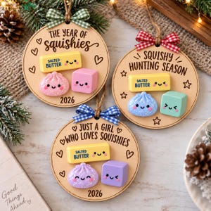 Könnte beinhalten: Drei runde Holzornamente mit Text und Miniatur-Squishy-Figuren. Eines zeigt THE YEAR OF SQUISHIES 2026, ein anderes SQUISHY HUNTING SEASON und das dritte JUST A GIRL WHO LOVES SQUISHIES 2026. Jedes hat eine kleine Schleife und eine Aufhängeschnur.
