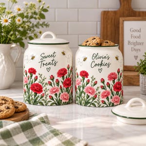 May include: Two cream-colored ceramic canisters with lids, decorated with red and pink carnation floral patterns, small bees, and black script text. One canister reads Sweet Treats and the other reads Olivia's Cookies, both featuring a small heart.