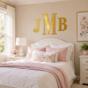 May include: Large gold-coloured wall letters J, M, and B mounted above a white bed frame. The bed features a white quilt, pink quilted pillows, and a rectangular accent pillow with a pink floral pattern. A white lamp sits on a bedside table.