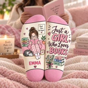 May include: A pair of cream-colored socks with pink toe and heel sections. The left sock features an illustration of a person reading with a cat, the name EMMA, and text reading Read More Be Happier. The right sock displays the text Just a GIRL Who Loves BOOKS with a bouquet of flowers and a stack of books labeled COZY BOOKS HAPPY LIFE.