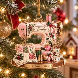 May include: A decorative Christmas ornament shaped like a pink sewing machine with gold accents. It features a quilt, spools of thread, scissors, a pincushion, and a thimble. The name Emily is printed on the machine, and the base reads Sew Create Belong.