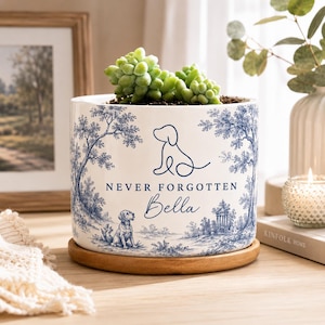 May include: A white ceramic cylindrical planter with a blue toile-style pattern featuring trees, a gazebo, and a dog. The text reads NEVER FORGOTTEN and Bella, accompanied by a line-art dog silhouette. It sits on a circular wooden saucer.