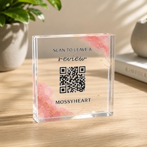 May include: A clear acrylic square sign featuring a black QR code, the text "SCAN TO LEAVE A review" and "MOSSYHEART" in black lettering. The design includes decorative pink watercolor-style accents with thin gold veining in the corners.