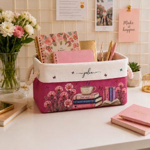 May include: A rectangular fabric storage bin with white rope handles, featuring a pink base with a floral and book graphic, a teacup illustration, and the text "Just one more chapter". The white top band is personalized with the name "julia".