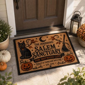 Peut inclure: Un paillasson rectangulaire en fibre de coco beige orné d'un chat noir, deux citrouilles d'Halloween, un balai et un croissant de lune. Le texte indique : THE JOHNSONS' SALEM SANCTUARY FOR WAYWARD CATS, FERALS & FAMILIARS WELCOME, EST. 1692.