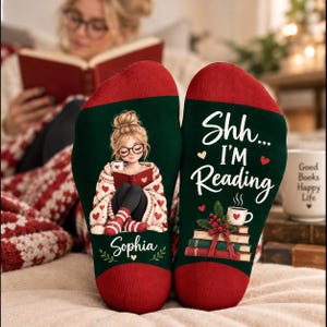 Può includere: Un paio di calzini verde scuro con punte e talloni rossi. Il calzino sinistro presenta l'illustrazione di una persona con gli occhiali che legge un libro, con il nome Sophia sotto. Il destro mostra il testo Shh... I'M Reading con una pila di libri e una tazza di caffè.