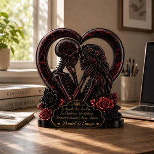 May include: A heart-shaped sculpture featuring two skeletons facing each other, adorned with red and black roses. A plaque at the base reads: In Death, We Are One. In Darkness, We Belong. Forever Entwined, Never Apart. Daniel & Emma.