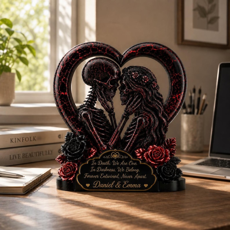 May include: A heart-shaped sculpture featuring two skeletons facing each other, adorned with red and black roses. A plaque at the base reads: In Death, We Are One. In Darkness, We Belong. Forever Entwined, Never Apart. Daniel & Emma.