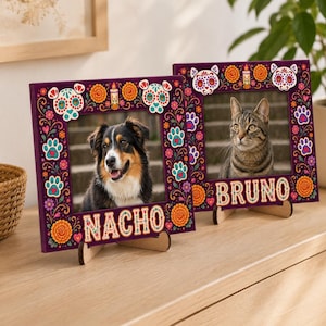 May include: Two square wooden picture frames with a dark purple background featuring colourful Day of the Dead inspired floral patterns, sugar skull cat designs, and paw prints. One frame displays a dog and the name NACHO, the other a cat and BRUNO.