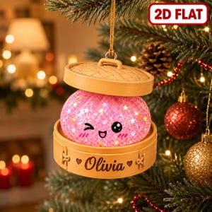 May include: A 2D flat Christmas tree ornament shaped like a bamboo steamer basket containing a pink, glittery, winking dumpling character. The base of the steamer is engraved with the name Olivia between two small heart icons.