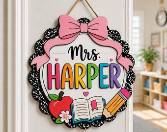 May include: A round wooden door sign featuring a black and white composition notebook pattern border. The center displays the text Mrs. HARPER in colorful letters, topped with a pink bow. It includes a red apple, an open book, and a yellow pencil.
