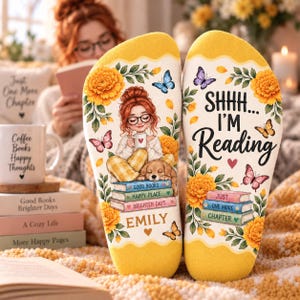 May include: A pair of cream-colored socks featuring a graphic of a person with red hair and glasses sitting with a dog on a stack of books. The text reads "SHHH... I'M READING", "GOOD BOOKS", "HAPPY PLACE", "BRIGHTER DAYS", "EMILY", and "JUST ONE MORE CHAPTER".