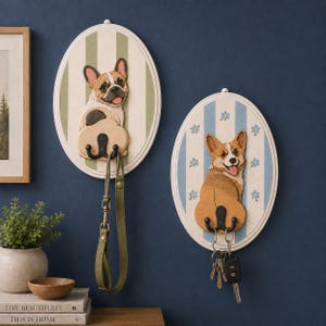May include: Two oval wall-mounted wooden plaques featuring carved dog figures. The left plaque has a French Bulldog on a green striped background with a black hook holding a leash. The right plaque has a Corgi on a blue striped background with floral accents and a black hook holding a set of keys.