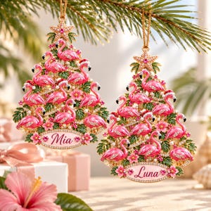 Pode incluir: Dois enfeites triangulares para árvore de Natal com flamingos cor-de-rosa, folhas de palmeira verdes e pequenas pérolas brancas. Cada enfeite é rematado com uma estrela dourada e tem uma base oval personalizada com os nomes Mia e Luna.