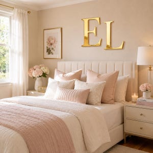 Könnte beinhalten: Große goldfarbene Metallbuchstaben E und L an einer beigen Wand über einem Bett. Das Bett verfügt über ein weißes, gestepptes Kopfteil, eine weiße Bettdecke, einen strukturierten rosa Bettläufer und mehrere Zierkissen in Weiß und zartem Rosa.