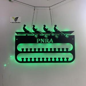 Puede incluir: Expositor de medallas de pared de metal negro con una silueta de un equipo de remo en un bote. El expositor tiene dos filas de ganchos para medallas y el texto "PNRA" en el centro.