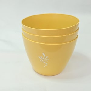May include: A stack of three smooth, yellow bowls with a tapered shape. Each bowl features a small, white, stylised floral or leaf-like graphic printed on the front centre. The bowls have a glossy finish and a simple, rounded rim design.