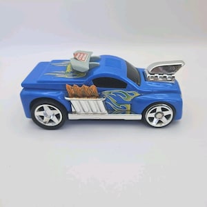 Puede incluir: Camión de juguete azul con llamas y un tubo de escape plateado. El camión tiene una parrilla plateada y una cama plateada con llamas naranjas. El camión es un juguete Hot Wheels.
