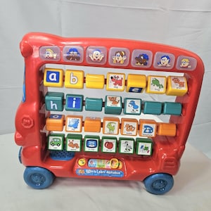 May include: A red plastic Spin & Learn Alphabus toy featuring four rows of rotating letter and picture blocks. The top row has character buttons, while the lower rows display letters like a, b, e, h, i, o and images of a dog, frog, goat, kitten, lamb, pig, rabbit, snake, telephone, van, worm, x-ray, yo-yo, zebra.
