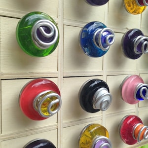 May include: A collection of 12 colorful glass drawer pulls with metal accents. The pulls are arranged in a grid pattern on a wooden surface. Each pull features a different color and design, including green, blue, red, yellow, and purple.