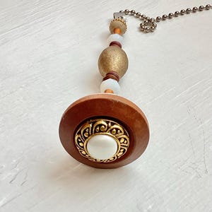 May include: A decorative ceiling fan pull chain with a wooden base and a gold-colored, ornate center. The chain features a series of beads in gold, white, and brown tones, connected by a metal ball chain.