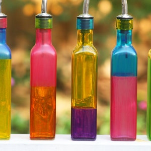 Pode incluir: Cinco garrafas de vidro com líquidos de diferentes cores dentro. As garrafas são todas do mesmo tamanho e formato, e têm uma tampa prateada no topo. Os líquidos são amarelos, laranja, roxos, rosa e verdes.