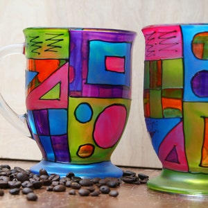Pode incluir: Duas canecas de vidro com desenhos geom&eacute;tricos coloridos pintados sobre elas. As canecas s&atilde;o ambas em um estilo semelhante, com uma mistura de quadrados, c&iacute;rculos e tri&acirc;ngulos em tons de rosa, azul, verde, roxo e laranja.
