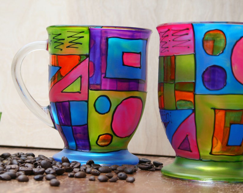 Pode incluir: Duas canecas de vidro com desenhos geom&eacute;tricos coloridos pintados sobre elas. As canecas s&atilde;o ambas em um estilo semelhante, com uma mistura de quadrados, c&iacute;rculos e tri&acirc;ngulos em tons de rosa, azul, verde, roxo e laranja.