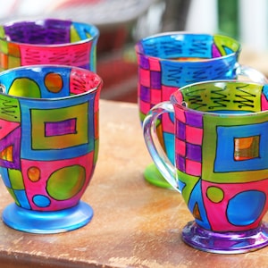 Pode incluir: Quatro canecas de vidro coloridas com desenhos geom&eacute;tricos pintados nelas. As canecas s&atilde;o de cores diferentes e t&ecirc;m padr&otilde;es diferentes. Elas est&atilde;o sobre uma mesa de madeira.