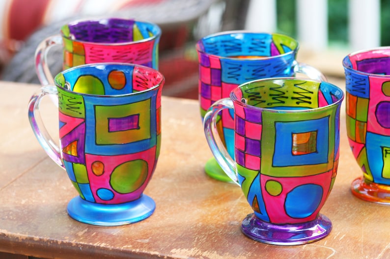 Pode incluir: Quatro canecas de vidro coloridas com desenhos geom&eacute;tricos pintados nelas. As canecas s&atilde;o de cores diferentes e t&ecirc;m padr&otilde;es diferentes. Elas est&atilde;o sobre uma mesa de madeira.