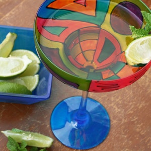 May include: A colorful glass margarita glass with a blue base and a painted design of geometric shapes in red, orange, yellow, green, and blue. The glass is filled with a clear liquid and garnished with a lime wedge and fresh mint.